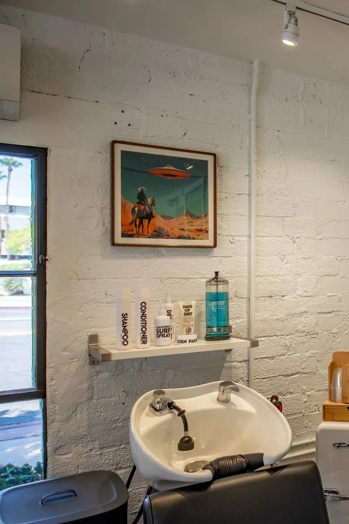 A vintage hair salon sink with a white brick wall in the background, displaying shelves with hair care products, a framed alien landscape artwork, and a window showing trees outside.
