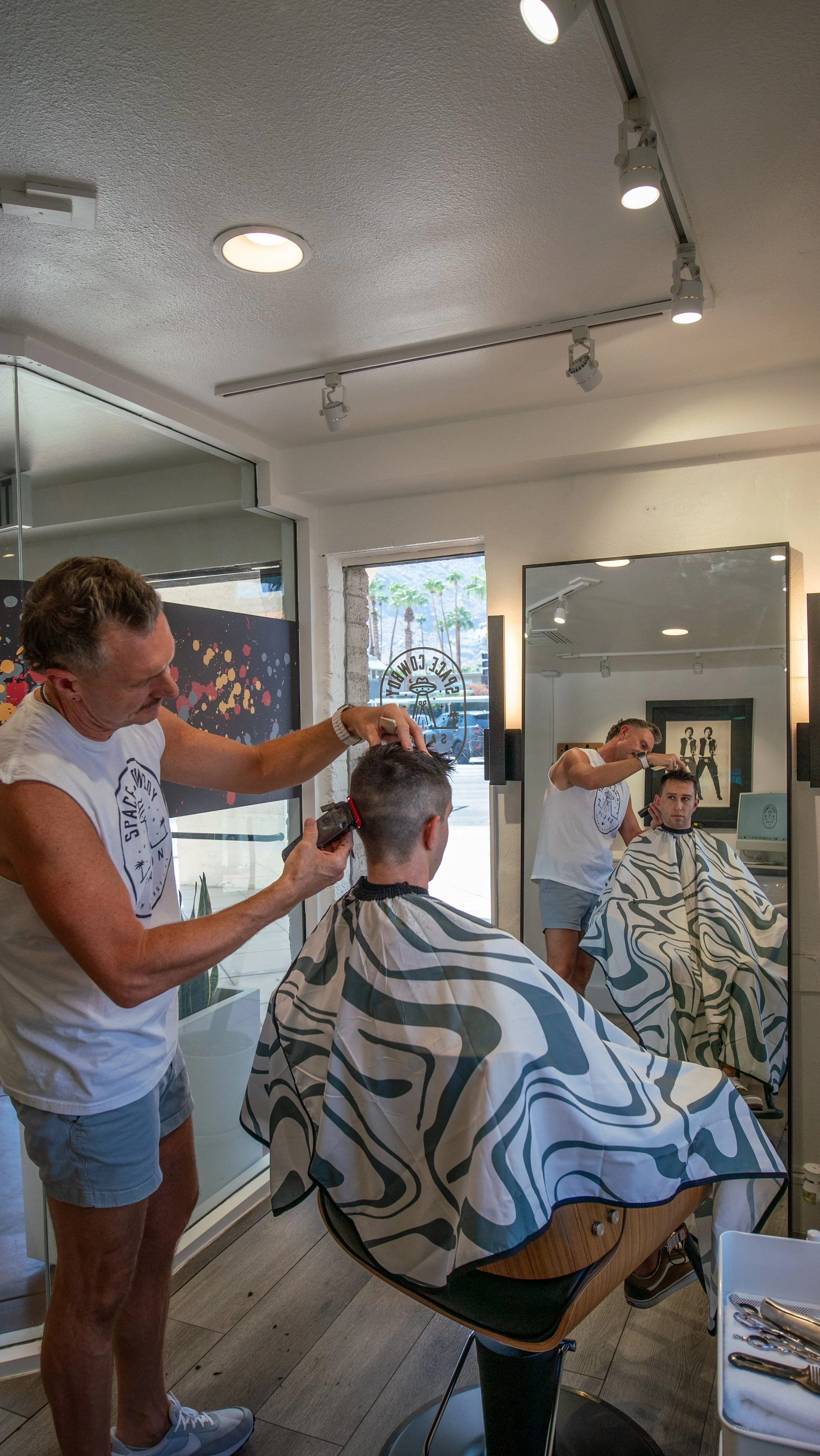 A man getting a haircut at a barbershop, seated in a chair covered with a black and white patterned cape, while a barber trims his hair with clippers. The barbershop has a large mirror, office decor, and bright lighting.