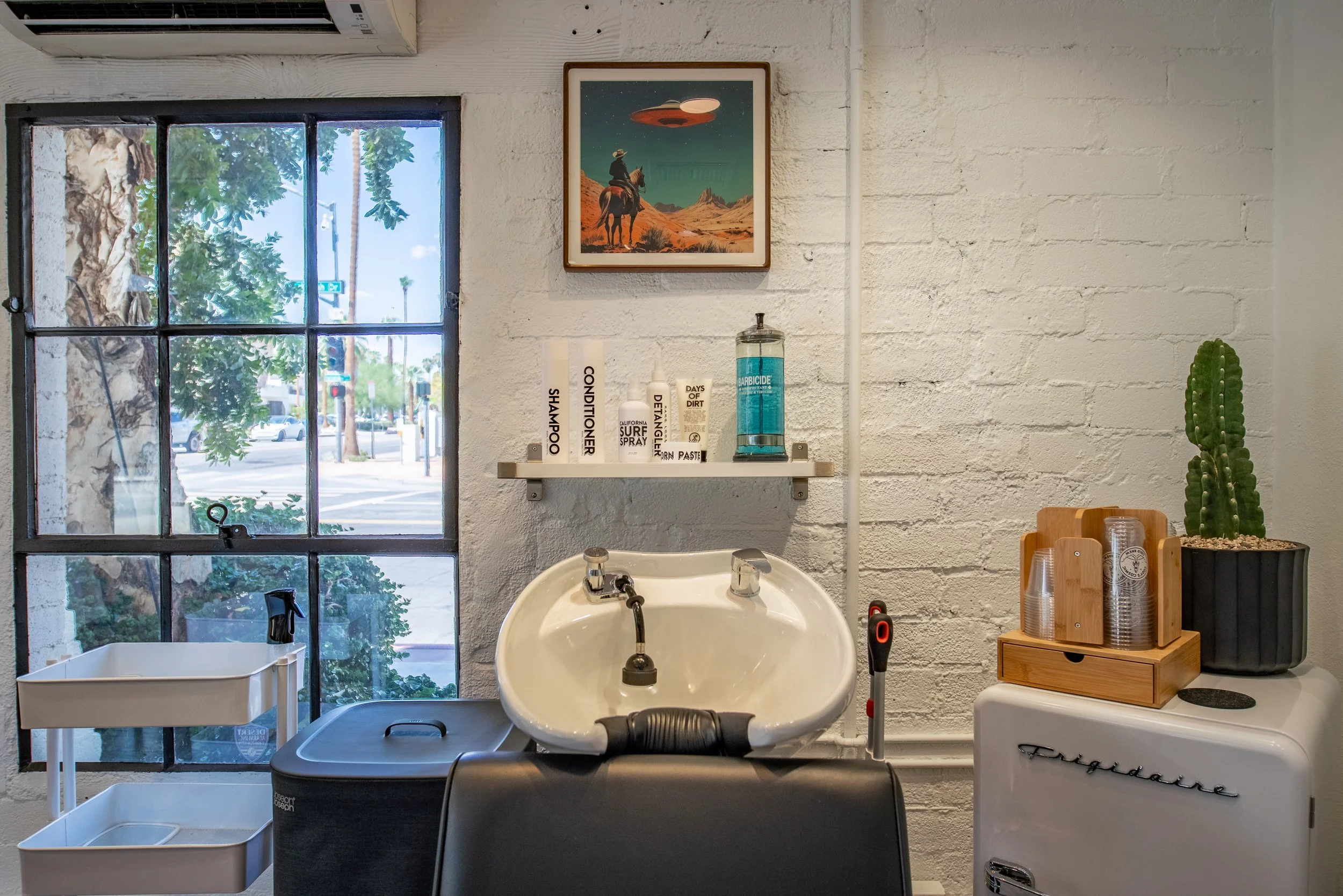 Inside a hair salon with a vintage-style white Frigidaire refrigerator, a large cactus in a black pot, a wooden caddy with disposable cups, a small white sink, a white brick wall, and a window showing street view with trees and traffic lights.