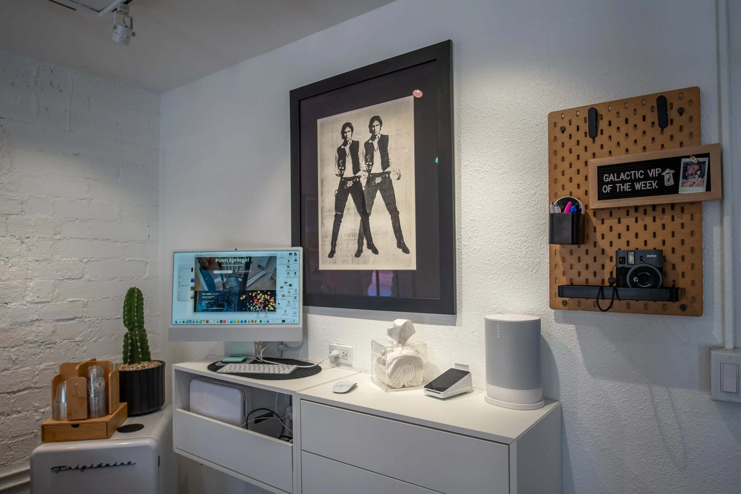Home office corner with a white desk holding a computer, tissue box, speaker, and phone. Above is a framed artwork of two women with short hair and sunglasses. On the wall to the right, a cork board with a sign reading 'Galactic VIP of the Week,' some pens, a small photo frame, a camera, and a container for supplies.