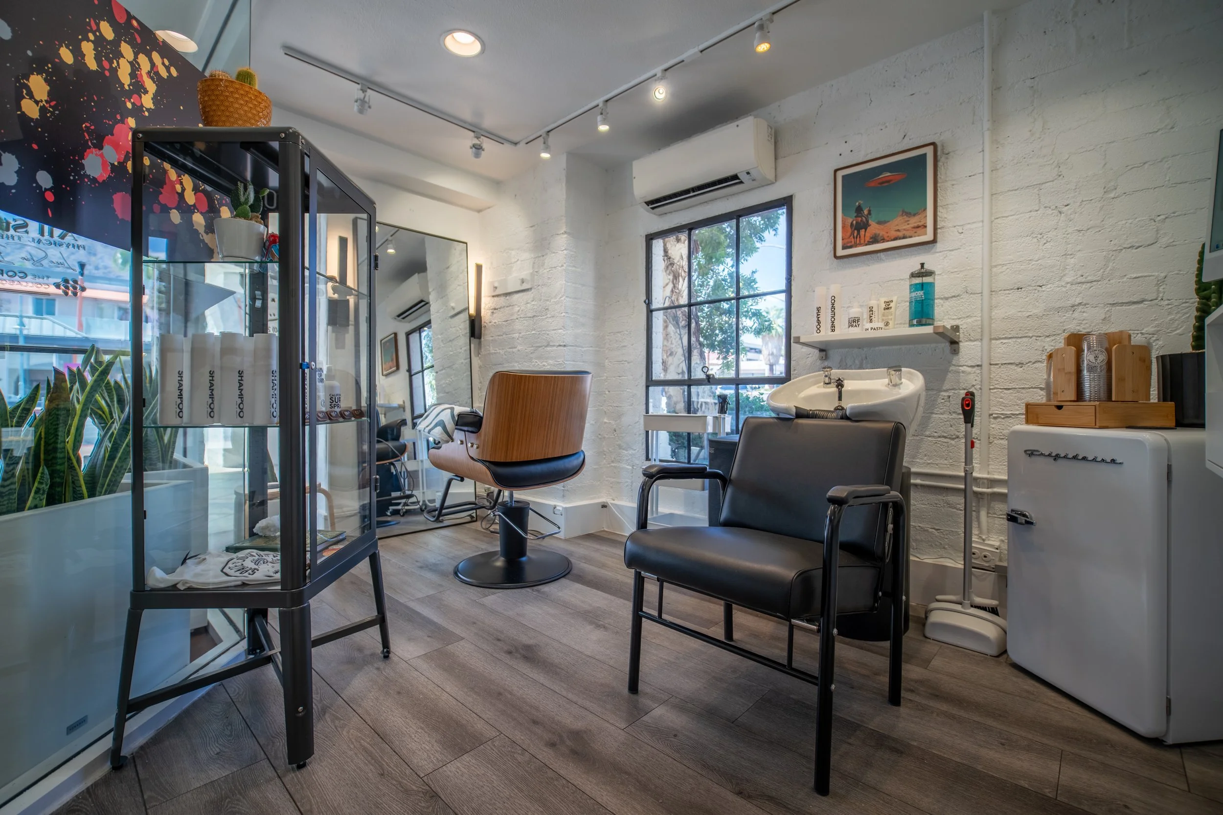 Interior of a modern hair salon with a styling chair, a wash basin, nail polish display, and decorative plants and art on white brick walls.