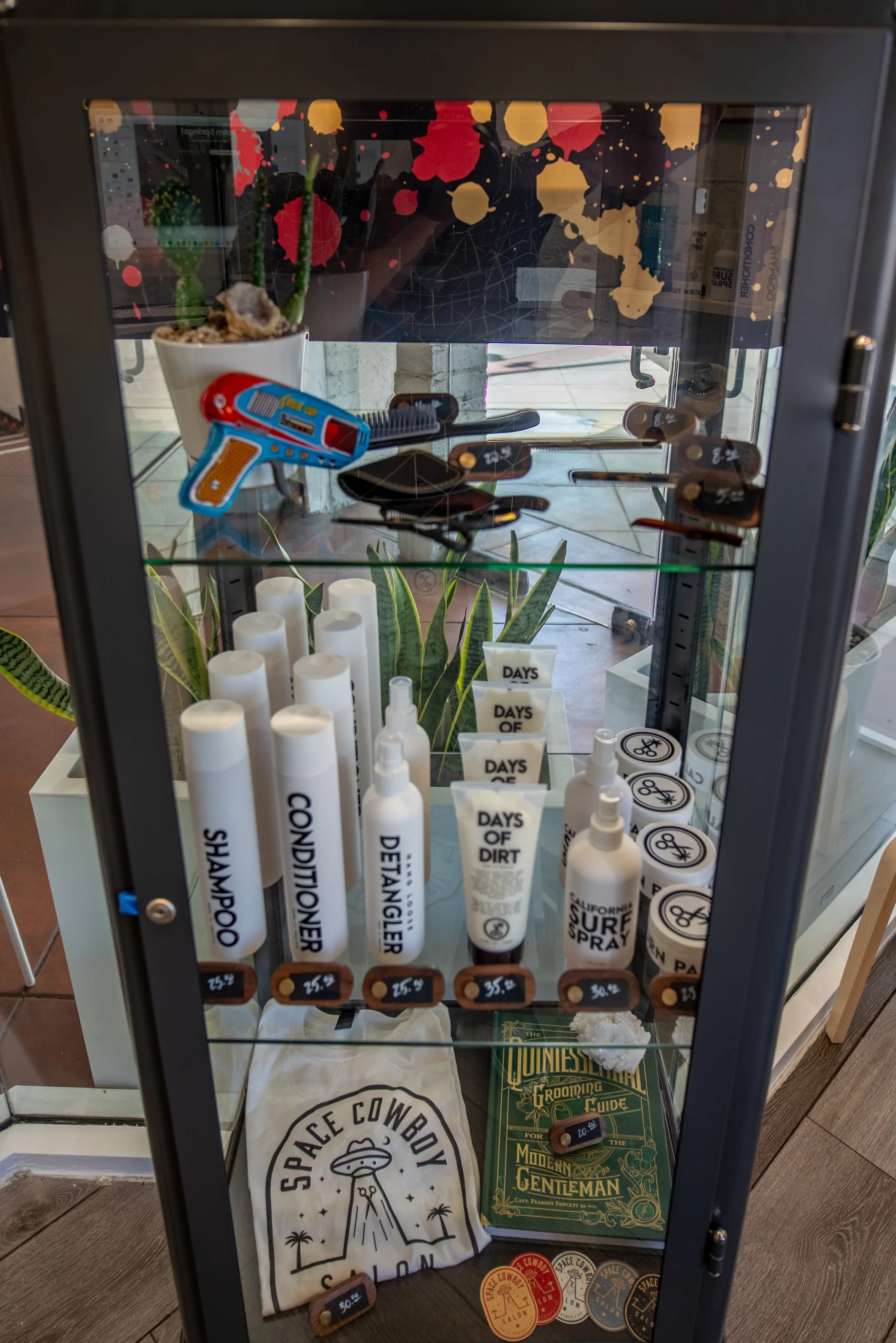 A display cabinet containing various grooming and skincare products labeled as shampoo, conditioner, deangeler, and dirt in white bottles, along with a plant and other small items inside.