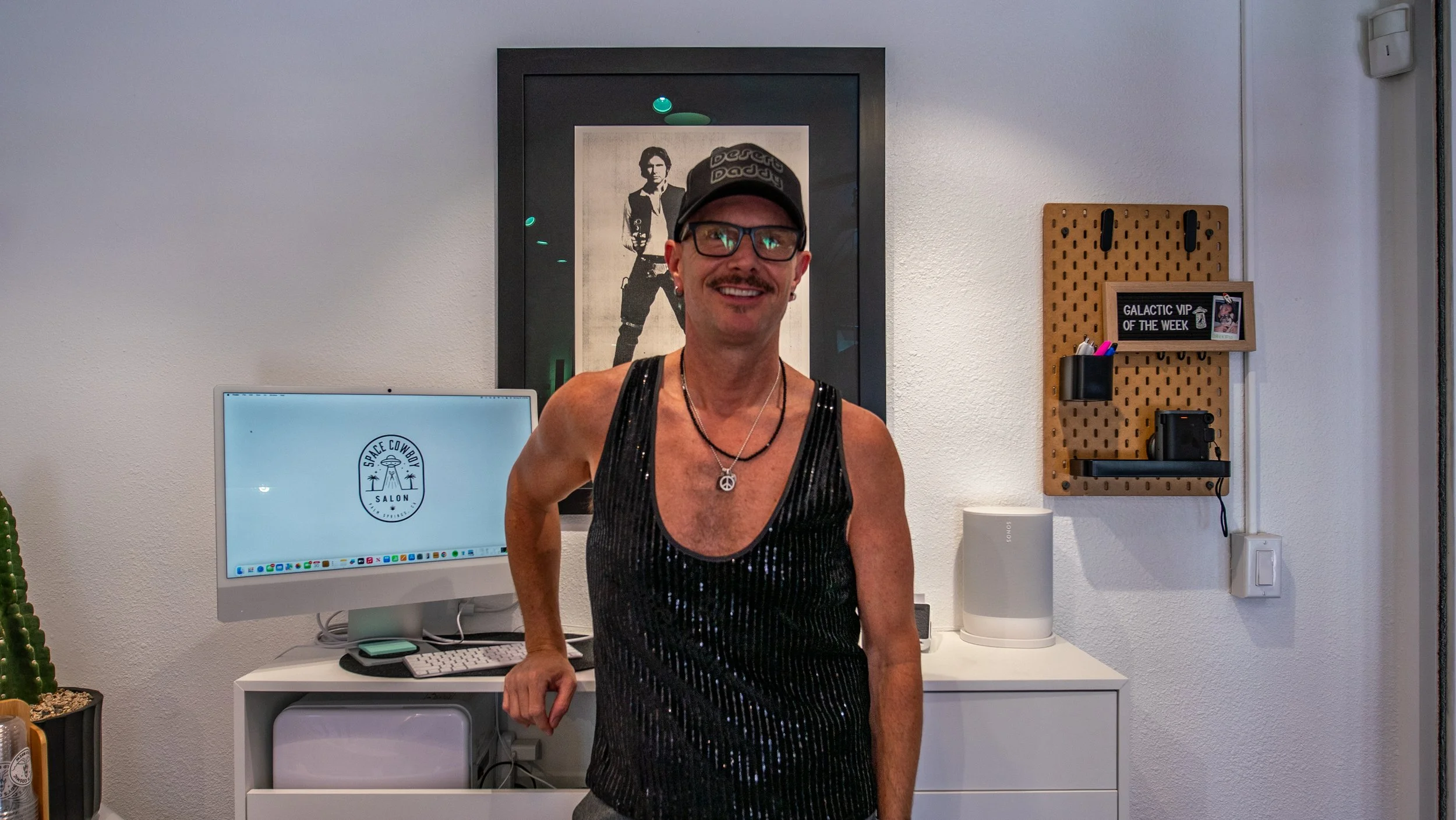 A man with glasses and a black cap smiling, wearing a black sequin tank top, standing in a modern office or salon with a framed photo of Elvis Presley in the background, a computer monitor displaying a logo, and a wall-mounted pegboard with a small framed sign, pens, a camera, and an electronic device.