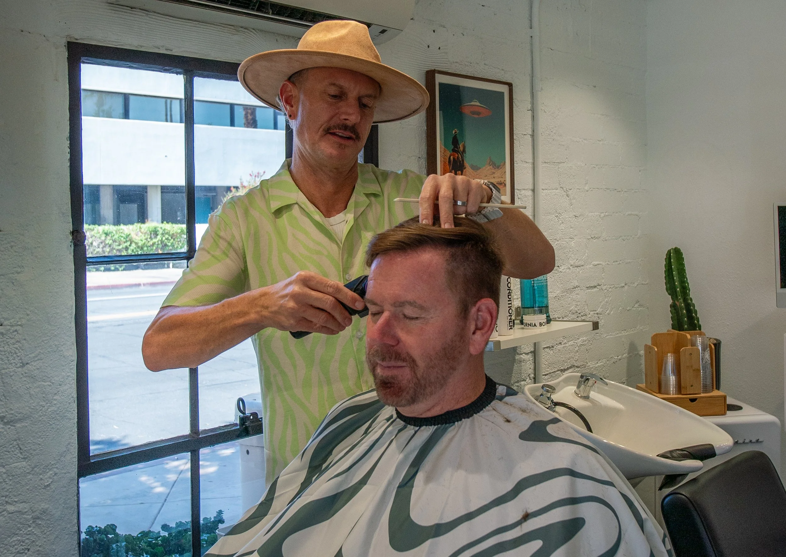 A man with short hair and a beard sits in a barber chair with a cape around his shoulders while a barber trims his hair using scissors and a comb inside a salon with white brick walls, a cactus, and art on the wall.